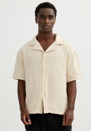 Hombre de pie mirando hacia adelante, vistiendo una camisa beige de manga corta con botones y cuello relajado sobre un fondo liso.