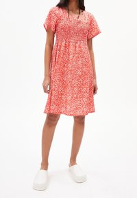 Krátké červené květinové šaty se smockovaným živůtkem, krátkými rukávy a splývavou sukní. Doplněné o bílé dřeváky na jednobarevném pozadí.