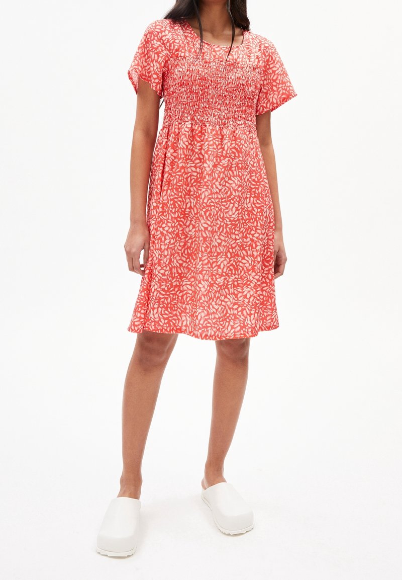 Krátké červené květinové šaty se smockovaným živůtkem, krátkými rukávy a splývavou sukní. Doplněné o bílé dřeváky na jednobarevném pozadí.