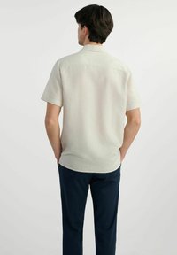 Krótki beżowy rękaw z luźnym fasonem, wykonany z lekkiego materiału. Posiada klasyczny kołnierzyk i wzór z tylnej części. Uzupełniony ciemnymi spodniami.