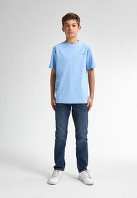 Camiseta de algodón de color azul claro con cuello redondo y mangas cortas, que presenta un diseño gráfico en azul más oscuro en el lado izquierdo, combinada con vaqueros oscuros.