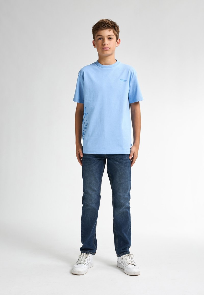 Camiseta de algodón de color azul claro con cuello redondo y mangas cortas, que presenta un diseño gráfico en azul más oscuro en el lado izquierdo, combinada con vaqueros oscuros.