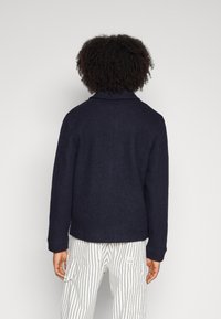 Navy wool blend jacket with a folded collar and long sleeves, paired with white striped pants. Features a textured finish.