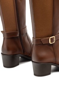 Botas marrones de cuero hasta la rodilla con un panel texturizado marrón, cremallera lateral y un adorno de hebilla en una correa; cuentan con un tacón bajo y grueso.