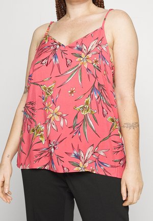 Donna che indossa un top smanicato floreale rosa con spalline sottili e pantaloni neri, mostrando tatuaggi su entrambi gli avambracci.