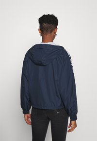 Person with short curly hair wearing hoop earrings, navy hooded jacket with text on the sleeve, and black jeans, facing away from the camera.