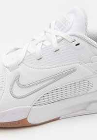 Bijela atletska cipela s teksturiranim mrežnim panelima, svijetlosivim akcentima i vidljivim Nike swoosh logom. Ima ravne vezice i gumeni potplat.
