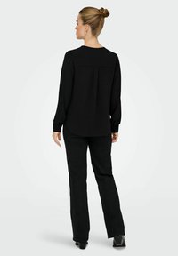 Haut noir à manches longues avec un col rond, détail plissé au dos, associé à un pantalon noir à jambes droites et des chaussures à talons.