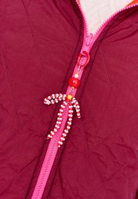 Czerwona pikowana kurtka z różowym suwakiem, z czerwoną i białą koralikową zawieszką do suwaka w kształcie małej postaci.