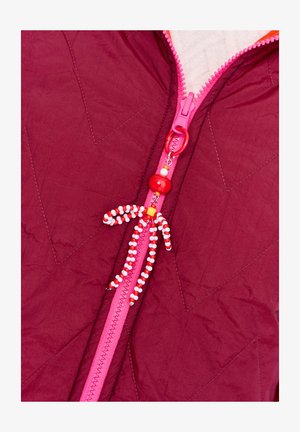 Czerwona pikowana kurtka z różowym suwakiem, z czerwoną i białą koralikową zawieszką do suwaka w kształcie małej postaci.