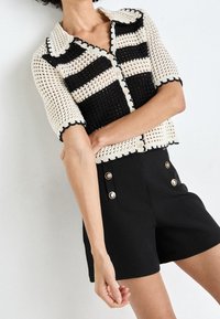 Kobieta ubrana w czarne szorty z wysokim stanem z złotymi guzikami oraz czarno-białą koronkową koszulę z krótkim rękawem, zapinaną na guziki, z kołnierzykiem.
