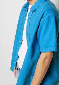 Camicia a maniche corte con bottoni in tessuto blu brillante, dal taglio rilassato, con bottoni dorati e design del colletto classico.