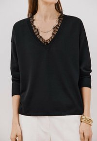 Maglione nero a maglia con scollo a V decorato con bordino in pizzo trasparente. Presenta maniche lunghe e una vestibilità comoda. Indossato sopra una collana a strati.