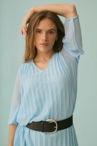 Femme aux longs cheveux bruns portant une robe plissée bleu clair et une ceinture noire, posant avec un bras levé sur un fond bleu pâle.