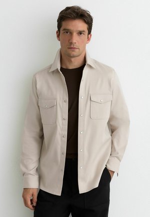 Mand iført beige skjorte med knapper over mørkebrun T-shirt og sorte bukser, stående mod en hvid væg med hænderne i lommerne.