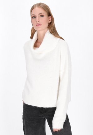 Eine Frau trägt einen weißen, flauschigen Rollkragenpullover und schwarze Hosen, steht vor einem schlichten weißen Hintergrund und blickt leicht nach links.
