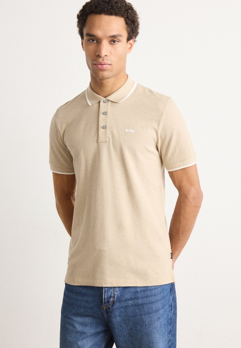Beige polo skjorte med korte ermer, hvit krage og mansjettstriper, pløs med tre knapper, subtil merking på brystet, glatt bomullstekstur.