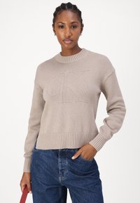 Maglione lavorato a maglia beige con scollo a girocollo, polsini a coste e un grande logo "CK" in rilievo sul davanti. Abbinato a jeans blu.