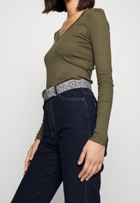 Olivgrünes, geripptes Langarmoberteil mit V-Ausschnitt, kombiniert mit dunkelblauen, hoch taillierten Jeans und einem schwarz-weiß gemusterten Gürtel.