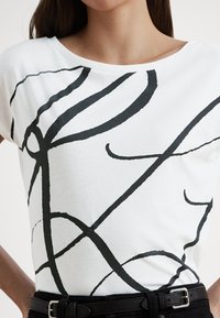 Balti medvilniniai marškinėliai su abstrakčiais juodais linijiniais raštais, plačiu kaklo iškirpimu ir laisvu siluetu. Derinami su tamsia juosmens juosta.