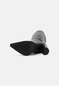 Botte à talon haut en matériau texturé gris avec un bout pointu et une semelle en caoutchouc noire présentant des lignes horizontales et des accents côtelés.