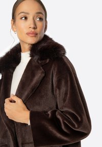 Abrigo de pelo sintético marrón con una textura suave, amplios solapas y un suave cuello de piel. Mangas largas y detalles de bolsillos visibles en la parte delantera.