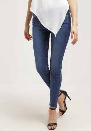 Mujer con jeans ajustados azules, sandalias negras de tacón alto y una blusa blanca asimétrica, de pie sobre un fondo liso.