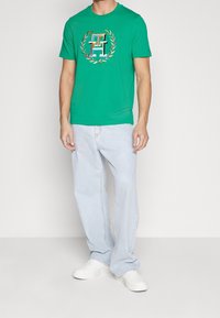 Camiseta de algodón verde con cuello redondo y un logo colorido rodeado por un diseño de laureles, combinada con jeans de mezclilla azul claro y zapatillas blancas.