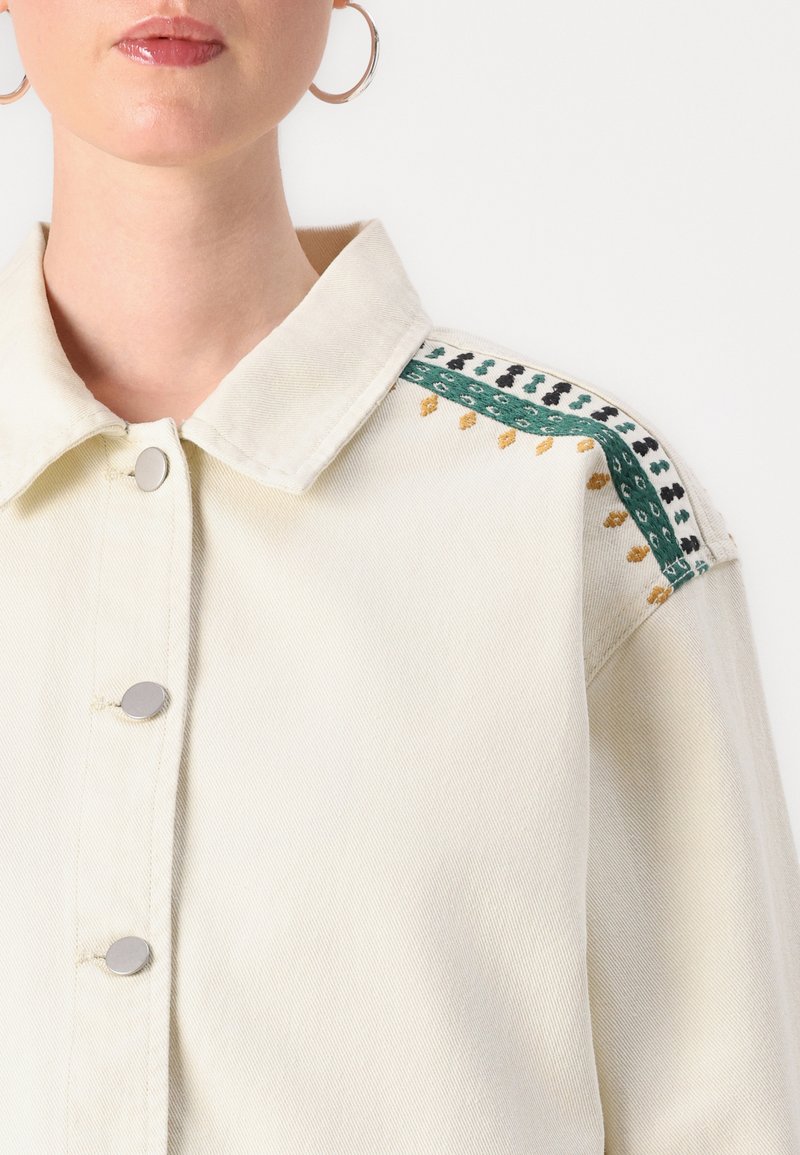 Veste en denim couleur crème avec un col, des boutons argentés et des motifs brodés décoratifs en vert, noir et or sur l'épaule.