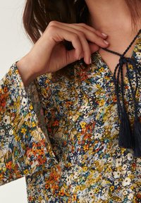 Blouse fleurie à manches longues, avec un motif multicolore de fleurs jaunes, rouges et bleues. Se noue au niveau du col avec des pampilles.