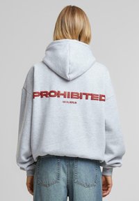 Sweat à capuche surdimensionné gris avec un cordon de serrage et des poches latérales. Il arbore le texte rouge "PROHIBÉ" et "10119, BERLIN" au dos.