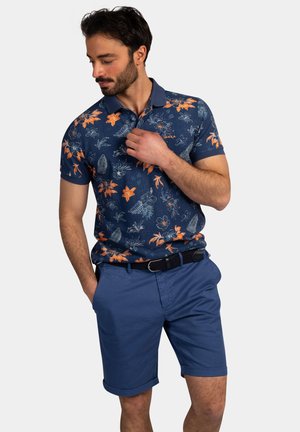 Man draagt een marineblauw poloshirt met oranje bloemen en blauwe korte broek, één hand in de zak, kijkt naar beneden en past de kraag aan.