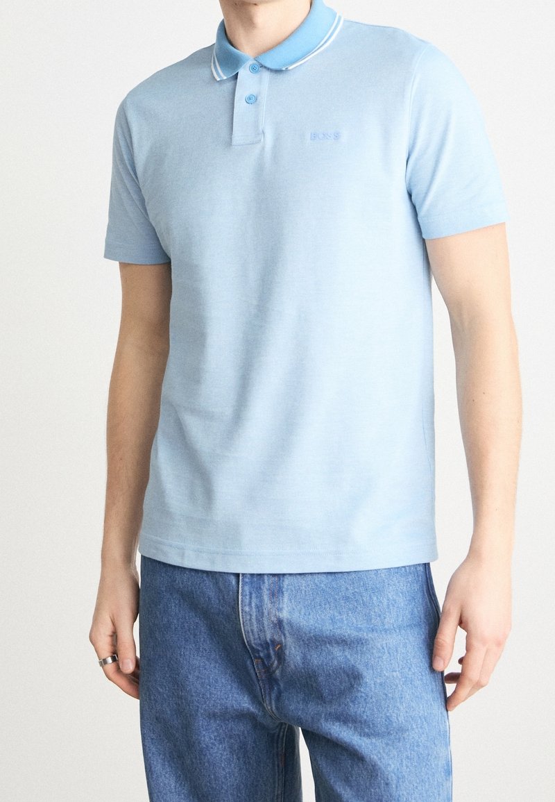 Homme portant un polo bleu clair à manches courtes avec col boutonné et un jean bleu, debout devant un fond uni.