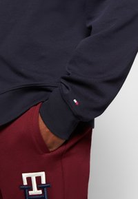 Felpa blu navy con polsini a coste, caratterizzata da un piccolo logo a forma di bandiera rossa, bianca e blu sulla manica. Pantaloni bordeaux con dettaglio ricamato bianco.
