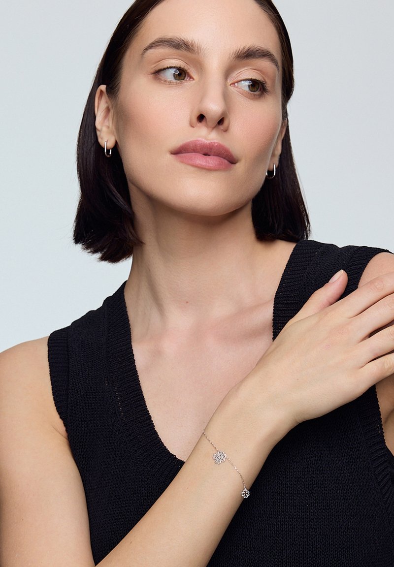 Srebrna bransoletka z kwiatowymi wzorami o skomplikowanych wycięciach, z delikatnym łańcuszkiem. Ubrana w czarny bezrękawnik z dzianiny, modelka pozuje pewnie.