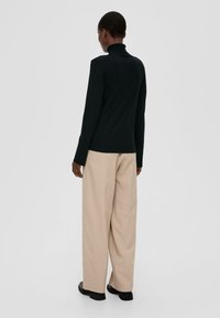 Mustahdoh svart turtleneck med långa ärmar, kombinerad med högmidjade beige vida byxor och svarta skor, sedd bakifrån.