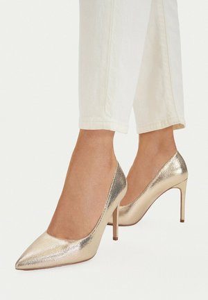Füße mit metallisch goldfarbenen spitzen High Heels kombiniert mit verkürzten weißen Hosen vor einfachem Hintergrund.