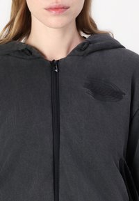 Sudadera con capucha gris con cremallera y acabado texturizado, con capucha ajustable con cordón y un logo sutil en el pecho. Cierre de cremallera y bolsillos delanteros.