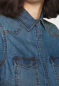 Denimskjorta i ljusblå färg, klassisk krage, två bröstfickor med knäppningar och kontrasterande sömmar.