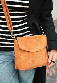 Frau in einem schwarz-weiß gestreiften Shirt und schwarzem Mantel, die eine beigefarbene Wildleder-Crossbody-Tasche mit einem Ledertrageriemen trägt.