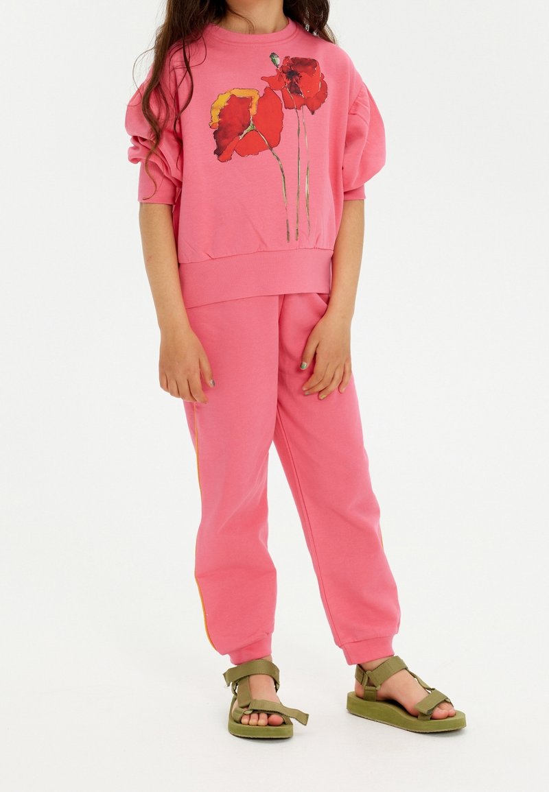 Enfant portant un pantalon de survêtement rose et un sweat-shirt avec des fleurs de coquelicot rouges, associé à des sandales vertes, debout sur fond blanc.