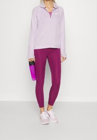 Top sportivo zip-up di un viola chiaro con colletto arrotondato, abbinato a leggins fitted di un viola scuro. Tiene una borraccia rosa trasparente.