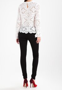 Valkoinen pitsipaita, jossa on kukkakuvioita ja pitkät hihat, yhdistettynä mustiin tiukkoihin farkkuihin sekä punaisiin ja vaaleanpunaisiin korkokenkiin. Selässä on avainaukkoyksityiskohta.