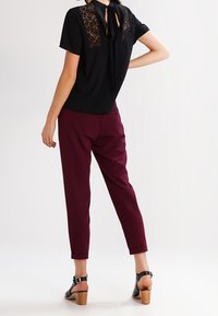 Camisa negra de manga corta con panel de encaje y detalle de lazo en la parte de atrás, combinada con pantalones burdeos y sandalias de tacón negras.