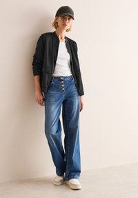 Veste bomber noire avec poches avant, t-shirt blanc, jeans larges bleus avec détails de boutons, et baskets blanches. Modèle portant une casquette verte.