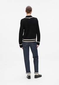 Schwarzer gerippter Pullover mit weißen Akzenten, Rückansicht. Kombiniert mit dunklen Jeans, hochgekrempelten Ärmeln und schwarzen Schuhen. Texturierte Stoffdetails.