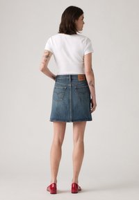 Falda de mezclilla, azul claro, diseño en A, dobladillo deshilachado, dos bolsillos traseros. Llevada con una blusa ajustada blanca y zapatos rojos brillantes.