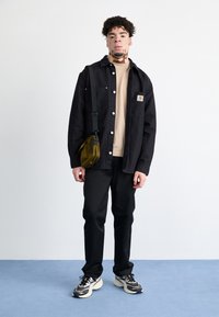 Sort knapjakke over en beige skjorte, mørke bukser og mønstrede sneakers. Olivengrøn crossbody taske. Neutral baggrund og blå gulv.