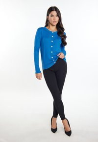 Cardigan blu a maniche lunghe con bottoni a forma di cuore, abbinato a leggings neri e tacchi alti neri, su uno sfondo bianco.