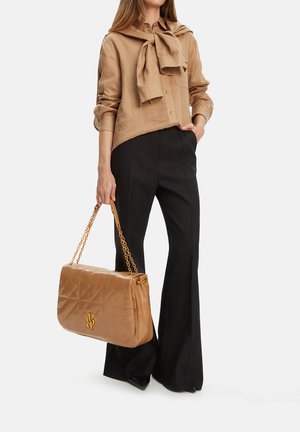 Žena oblečená v béžové košili na knoflíky s odpovídajícím svetrem přehozeným přes ramena, černé kalhoty, drží velkou béžovou prošívanou kabelku se zlatými řetízkovými popruhy.